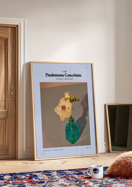 A framed Diadumene Leucolena poster featuring a beige flower in a vintage blue vase, set against a soft, neutral background, displayed in a cozy, well-lit room with a modern interior.