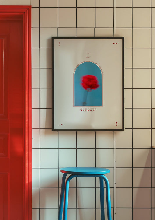 A minimalist poster featuring a blurred red flower in an arch design with the quote &quot;Love is fleeting, hold on to it&quot; in a modern, tiled bathroom. The poster is framed in black and adds a bold contrast to the white tiles and vibrant red door.