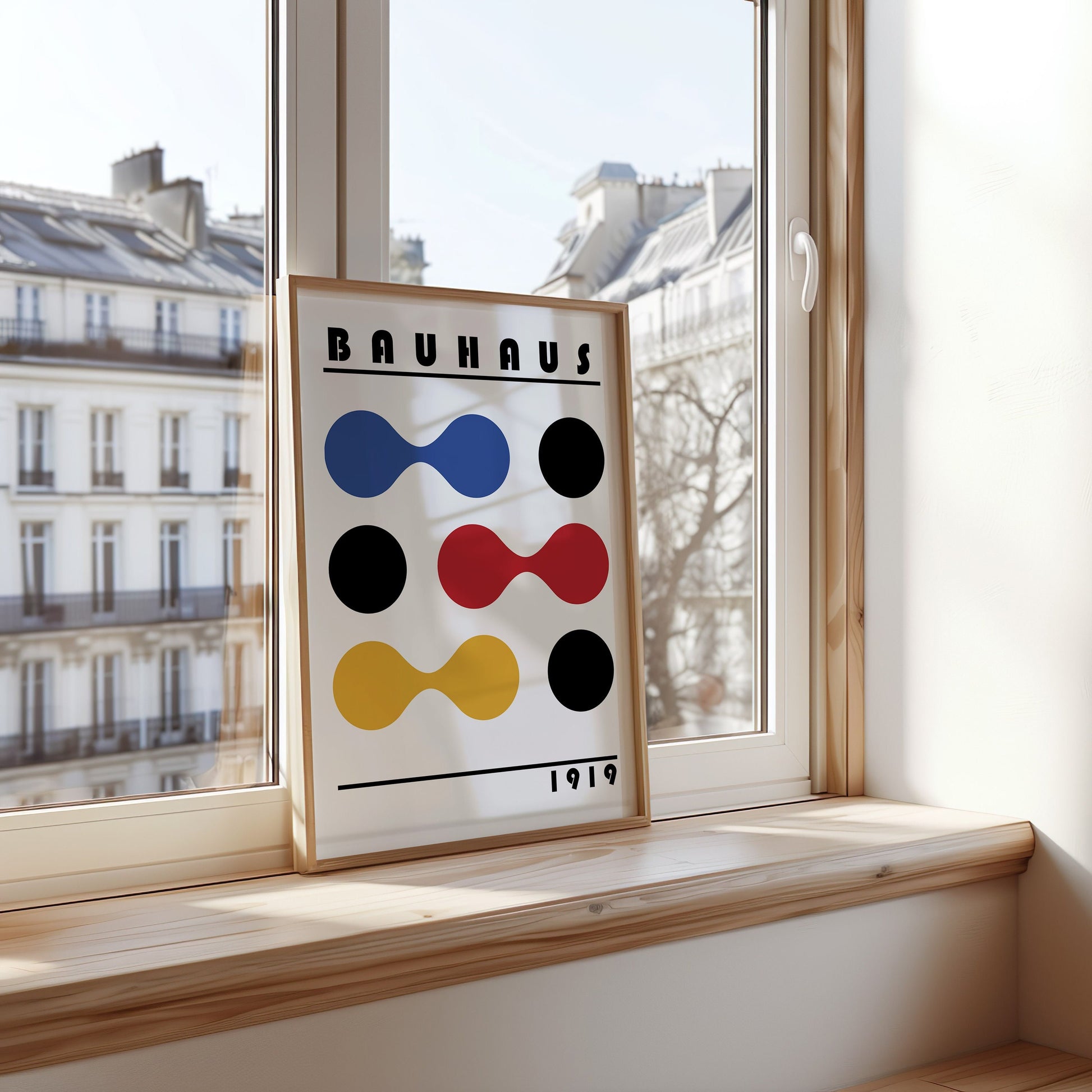 A Bauhaus poster from 1919 featuring a geometric design with primary colors (blue, red, yellow) and black circles, displayed on a light wooden frame by a window.