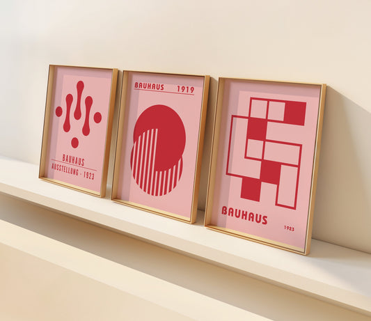 A set of three Bauhaus-inspired posters in bold red hues with geometric designs, framed in dark wood, displayed on a light wooden floor against a white wall.