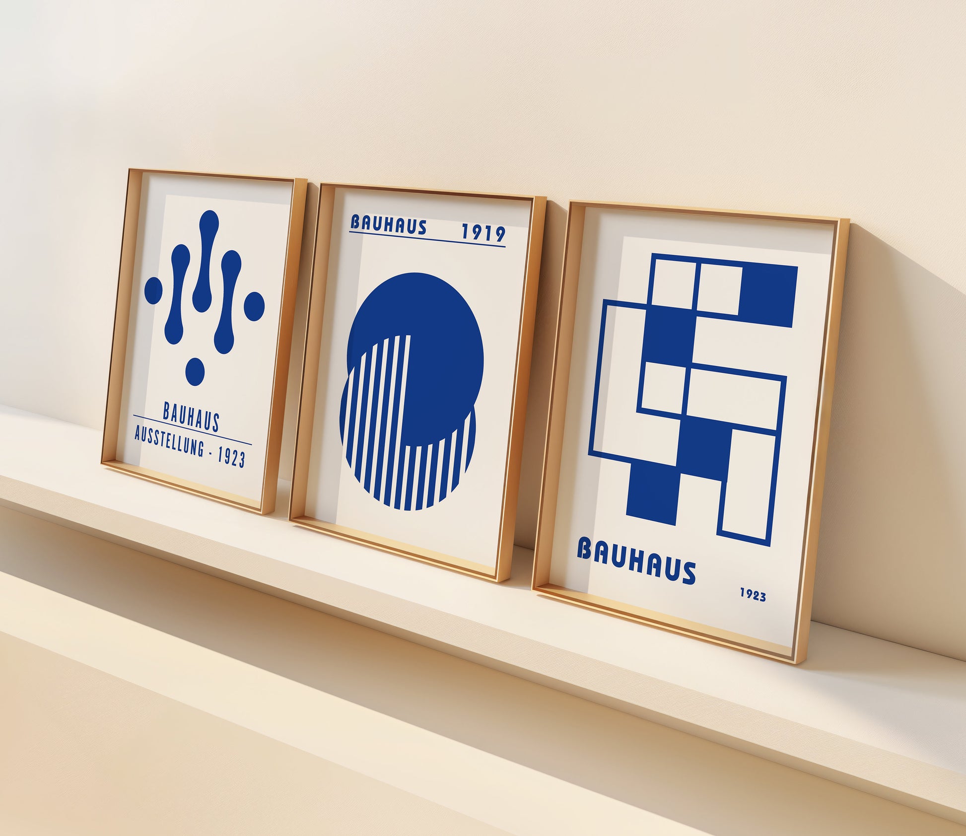 A set of three Bauhaus-inspired posters in vibrant blue hues with geometric designs, framed in dark wood, displayed on a light wooden floor against a white wall.