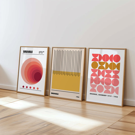 A set of three vibrant Bauhaus-inspired posters featuring abstract geometric designs in shades of red, yellow, and pink. The framed prints are beautifully displayed in a cozy, sunlit room with wicker furniture and light wooden elements.