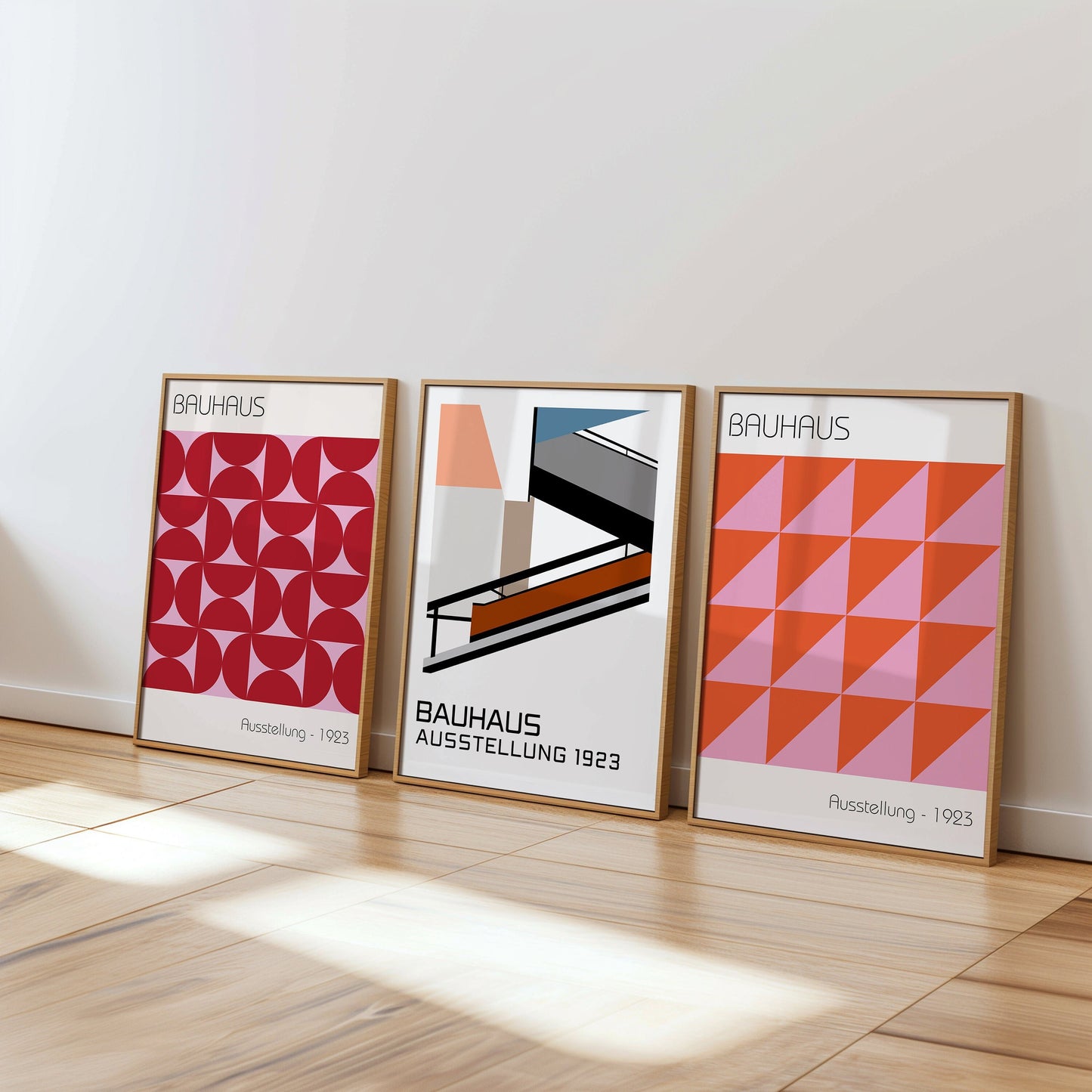 A set of three Bauhaus-inspired posters featuring abstract geometric designs, including triangular and curved patterns in red, pink, and orange. The posters are framed and displayed on a light wooden floor, creating a modern and artistic atmosphere.