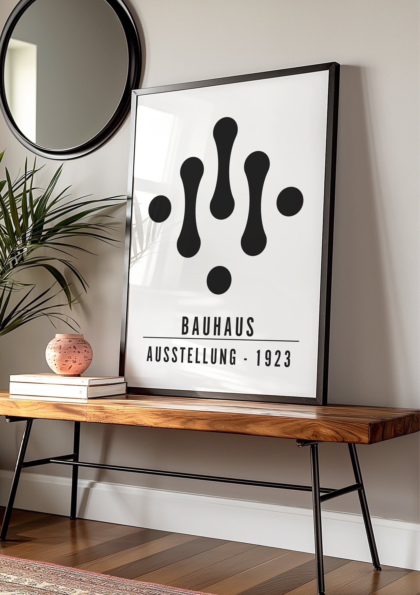 A framed Bauhaus Ausstellung 1923 poster featuring a minimalist black and white geometric design. The poster leans against a wall on a wooden bench in a modern living room, complemented by a round mirror and a potted plant.