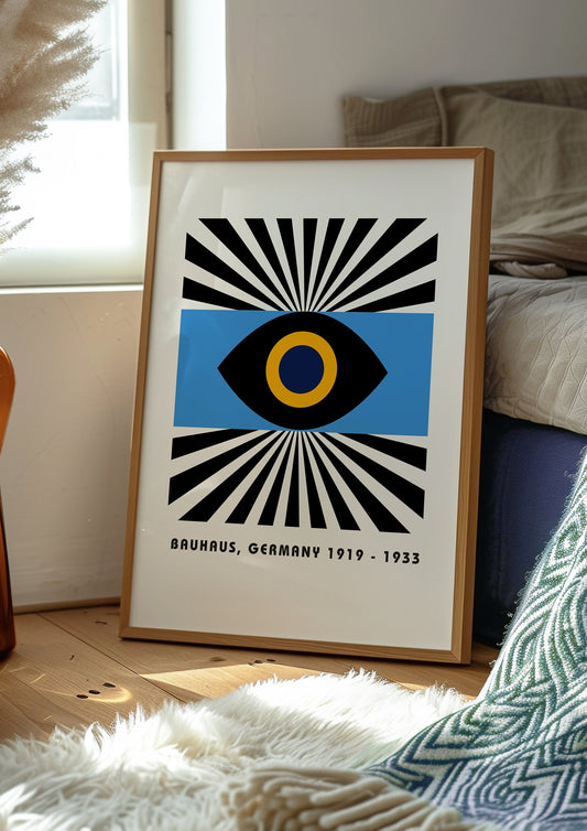 A striking Bauhaus-inspired poster featuring a geometric eye design in black and yellow against a blue background, framed by bold black and white rays, hanging on a tiled wall with a red door and blue stool nearby.