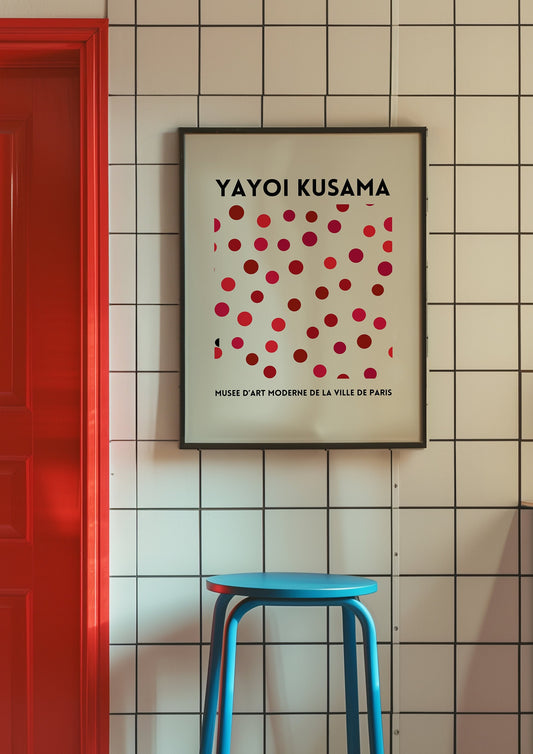 A framed Yayoi Kusama-inspired red dot poster, featuring a minimalist and abstract dot design in shades of pink and red, leaning against a rustic wall next to a pink chair.