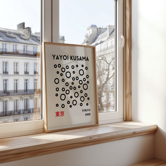 A framed Yayoi Kusama-inspired poster with black dots and a minimalist design featuring a red-haired figure peeking over the top. The print is set in a modern room with stylish decor.