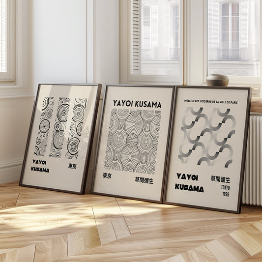 A set of three Yayoi Kusama posters featuring minimalist abstract circle patterns in black and white, displayed on a floor by a bright window.