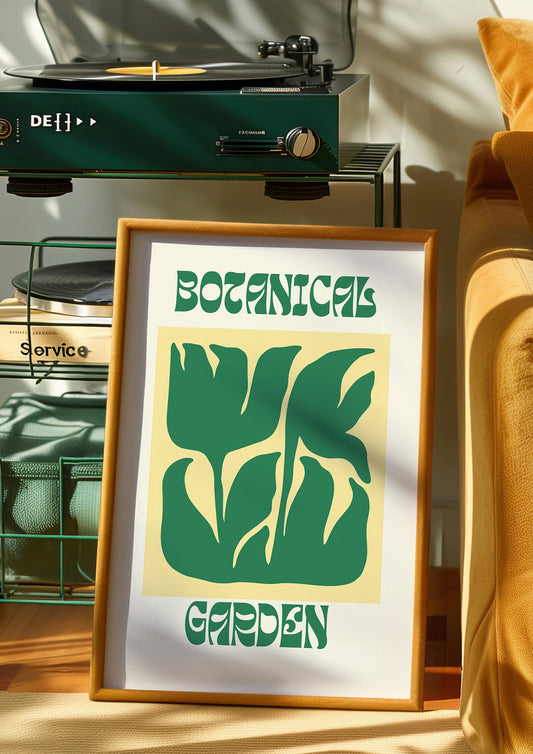 A &quot;Botanical Garden&quot; poster featuring a bold green leaf design on a soft yellow background, framed in gold and placed on a textured rug. The retro typography and abstract plant shapes evoke a mid-century modern aesthetic, perfect.