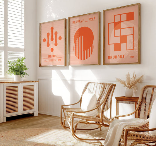 A set of three Bauhaus-inspired posters in vibrant orange hues with geometric designs, framed in light wood, displayed on a wooden floor against a white wall.