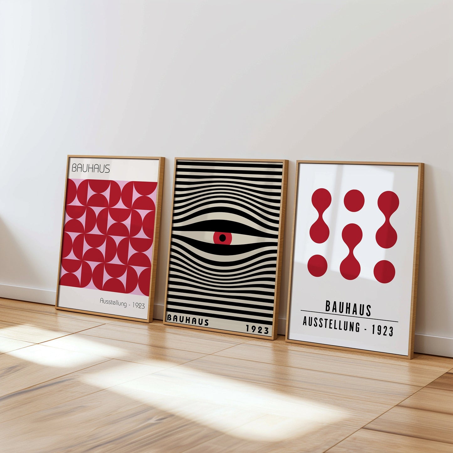 A stylish set of three Bauhaus posters featuring red and black geometric designs. The posters, framed in light wood, are displayed on a white wall in a modern living room with wicker chairs and a bright, airy atmosphere.