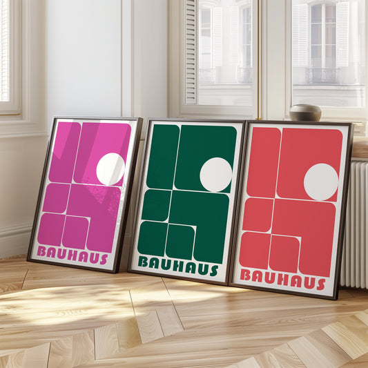 A vibrant set of three Bauhaus posters in pink, green, and red, featuring minimalist geometric designs with bold shapes. The framed prints are displayed on a wooden floor against a bright interior backdrop, adding a pop of color and artistic flair.