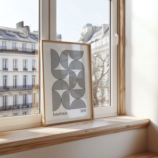 A framed Bauhaus poster with a black and white geometric pattern, leaning against a window in a modern apartment setting. The clean lines and minimalist design make it a perfect addition to any Scandinavian or contemporary decor.