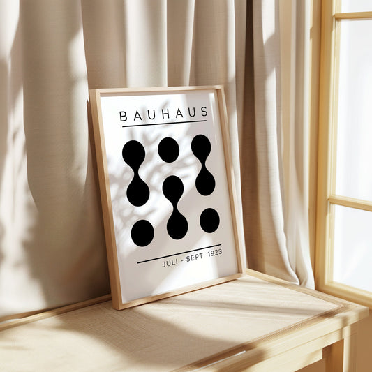 A Bauhaus exhibition poster from Juli-Sept 1923, featuring a minimalist black and white abstract design with geometric shapes, framed and leaning against a beige curtain in a softly lit room.