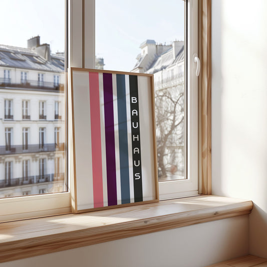 A minimalist Bauhaus-inspired art print featuring vertical stripes in shades of pink, purple, green, and blue with the word &quot;Bauhaus&quot; in bold white text, displayed by a window with a Parisian cityscape in the background.