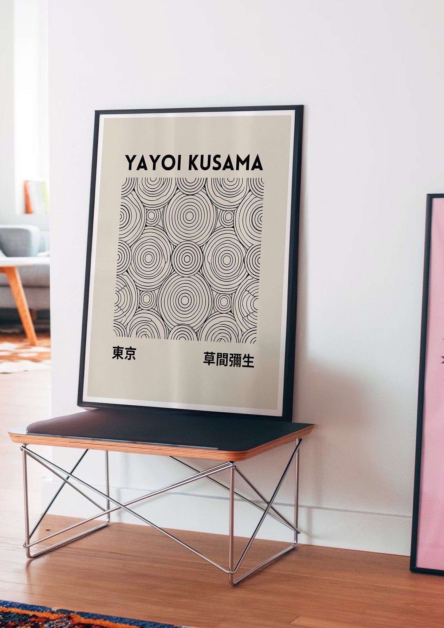 Framed Yayoi Kusama abstract circle print with black concentric circles on a beige background, propped against a white wall next to a modern bench. The minimalist design adds an elegant, contemporary touch to the interior.