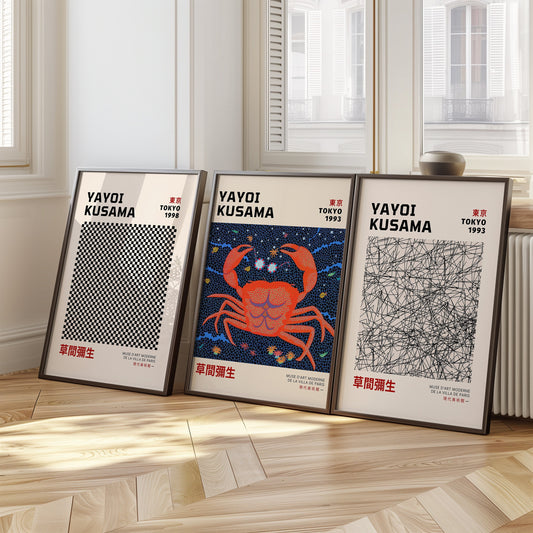 A set of three framed Yayoi Kusama posters displayed on a wooden floor. The first features a black and white geometric pattern from Tokyo 1998